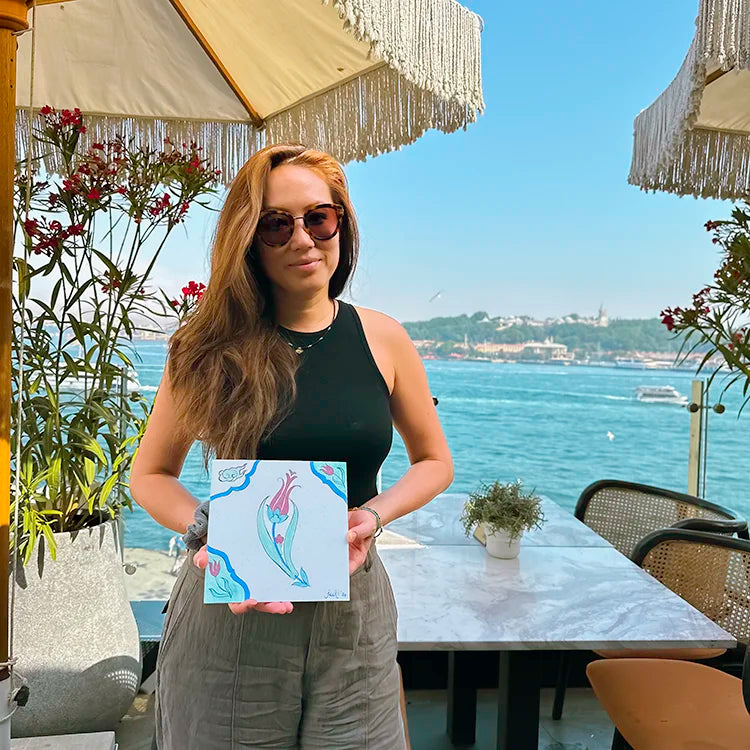 Young woman holding painted square tile, posing with the sea view on the background